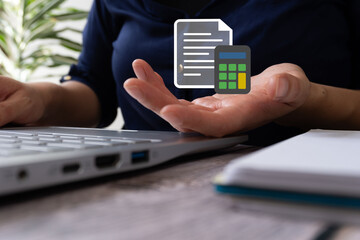Close-up of a woman's hand holding a digital calculator with a file document icon floating above, on a desk next to a laptop computer