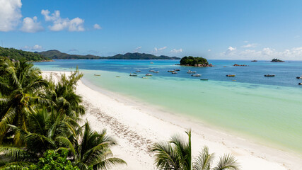 A pristine beach with soft white sand, lush palm trees, and boats floating in turquoise waters near a small island. Praslin, Seychelles.