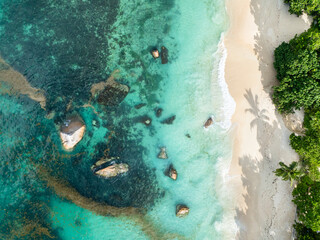 Clear turquoise waters reveal submerged rocks near a white sandy beach, with lush green trees...