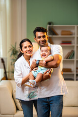 Young Indian parents standing with baby boy in arms inside modern stylish living room at home