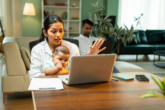 Mother working on laptop holding baby boy while father walks in background indoors at home