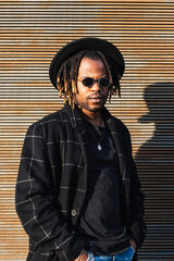 Modern adult black man with dreadlocks wearing hat, sunglasses and stylish coat looking at camera...