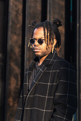 Frowning modern African American guy with dreadlocks hairstyle wearing sunglasses with coat...