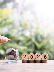 Hand putting coin into glass bottle, Wooden blocks with the number 2026 on stack of coins. The...