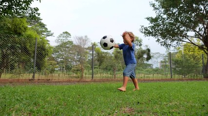 Barefoot young boy picks up soccer ball from grass, gently tosses it and makes volley pass as ball falls, sending it flying far. Child learning soccer skills shows very good ball control