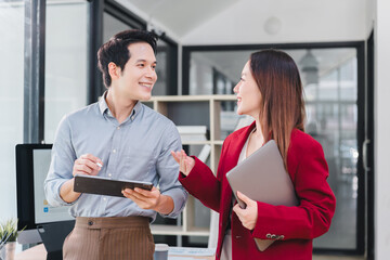 Smiling man and woman are having friendly conversation in modern office, holding tablet and laptop,...