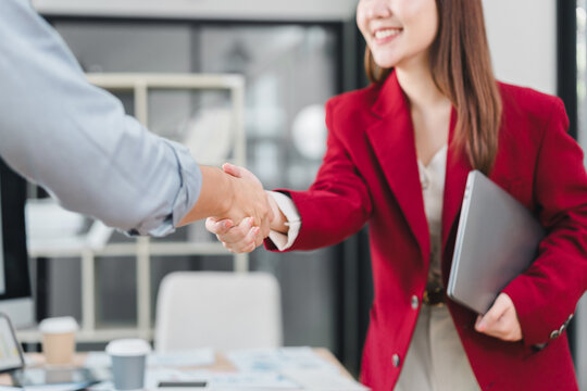 Two business people shaking hands in office, smiling and holding laptop, showing cooperation and agreement in professional environment - Powered by Adobe