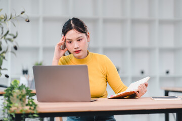 Young woman in yellow sweater working on laptop and holding book, showing focused and thoughtful expression in modern office environment