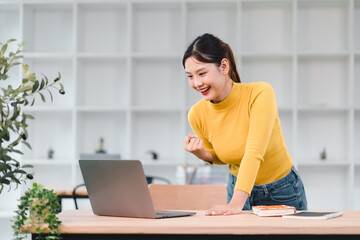 Young woman in yellow sweater smiles excitedly while looking at laptop on wooden table in bright modern room with plants and shelves in background