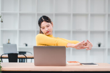 Obraz premium Young woman in yellow sweater stretches her arms while sitting at desk with laptop and notebooks, expressing tiredness or boredom in modern office setting