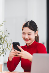 Young woman smiling while using smartphone indoors, wearing red sweater, sitting at desk with laptop and plant nearby, casual and modern setting