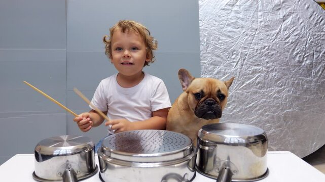 Adorable young boy enthusiastically plays makeshift drum set built from kitchenware, banging on steel pots with wooden sticks. Sitting nearby, his companion dog calmly endures noisy show