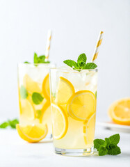 Two glasses of fresh lemonade with lemon slices, mint leaves, ice, and striped straws on a bright background