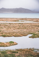Grass and stone shore of Lake Karakul with blue water and mountain peaks in the background, cloudy weather and sky with clouds and clouds in the Pamirs, panoramic landscape