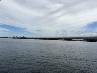Fototapeta premium Wide view of open water with distant factories or industrial buildings along the coastline under a cloudy sky.