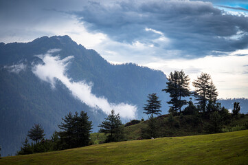 Mountain landscape with clouds.  At 6,900 feet,  serene meadows, pine forests, and proximity to the Great Himalayan National Park, Sainj Valley of Kullu District, Himachal Pradesh, India.
