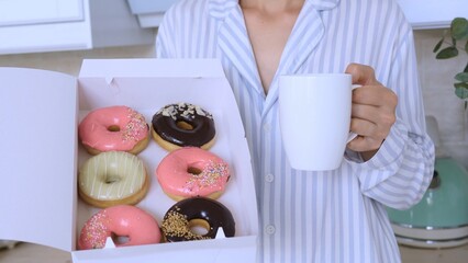 Woman wearing pajamas is holding a box of colorful donuts with various toppings and a white mug in...