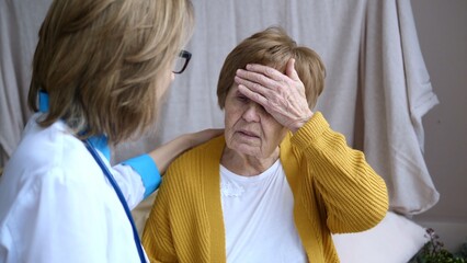 Female doctor is visiting and comforting a senior woman touching her forehead suffering from headache, dizziness, fever or migraine at home