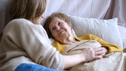 Young woman providing care for her sick elderly mother lying in bed, holding her hand and offering...