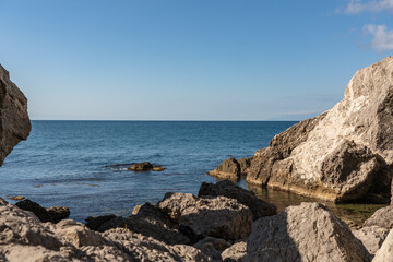 Coastal scene with clear blue water gently lapping against rugged rocks. Rocky coast of Black Sea. Cape Alchak. Sun casts warm glow, highlighting textures of stones and tranquil sea. Sudak, Crimea
