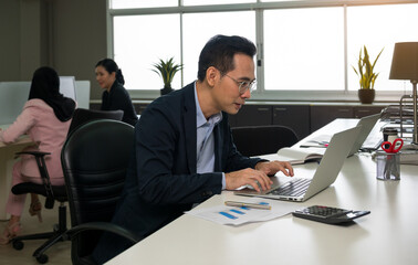 Professional businessman in dark suit working on laptop at modern office desk with documents and calculator. Colleagues visible in background near large windows.