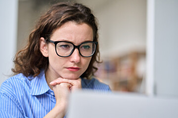 Busy young serious professional business woman employee or student wearing glasses using laptop watching online webinar or training web course, looking at computer, thinking, elearning. Close up