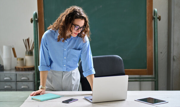 Young busy happy woman school professional teacher standing at desk in front of chalkboard in classroom working on laptop computer. Online tutor teaching remote education virtual classes. - Powered by Adobe