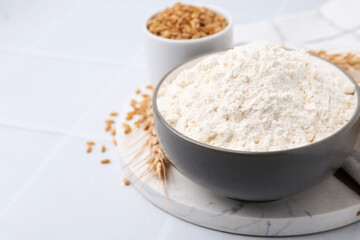 Wheat flour, grain and spikes on white tiled table, closeup. Space for text