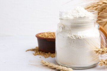 Wheat flour in glass jar, grains and spikes on white table, closeup. Space for text