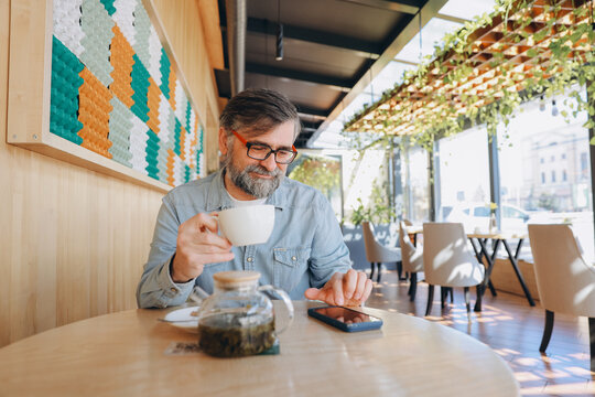 Senior man drinking tea and using smartphone in modern cafe