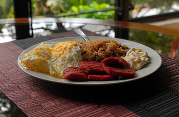 Gallo pinto, a typical Costa Rican dish with rice, red beans, sausage, cream, and a fried egg, and a glass of coffee on a table with a nature background.