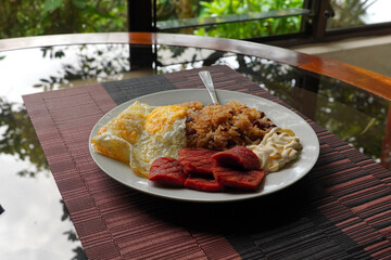 Gallo pinto, a typical Costa Rican dish with rice, red beans, sausage, cream, and a fried egg, and a glass of coffee on a table with a nature background.
