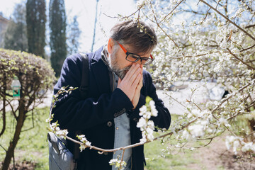 Senior man suffering from pollen allergy in spring next to blooming tree