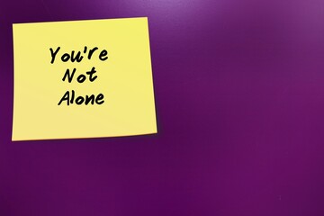 Stick note on purple copy space wall written YOU'RE NOT ALONE - positive self talk affirmation, encouragement message to support people when life seems difficult and hopeless