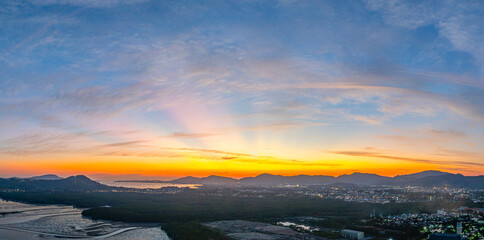 A stunning panoramic view of a coastal city at sunset at Phuket city, showcasing vibrant hues of orange and blue in the sky, with mountains, and urban lights creating a harmonious blend of natural