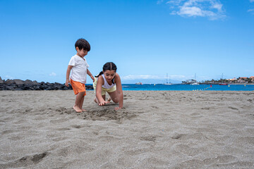 Two children playing on the beach. One is wearing an orange shirt and shorts. The other is wearing a white shirt