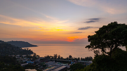  A stunning aerial view of the Andaman Sea at sunset, overlooking a coastal town in Phuket, Thailand. The golden hues of the sky reflect beautifully on the calm ocean waters, creating a tranquil