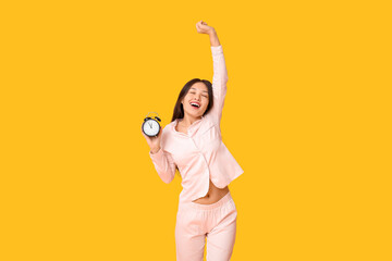 Beautiful young happy Asian woman in pajamas with alarm clock on yellow background