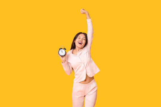 Fototapeta Beautiful young happy Asian woman in pajamas with alarm clock on yellow background