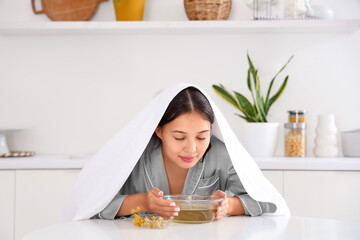 Young Asian woman doing steam inhalation at table in kitchen