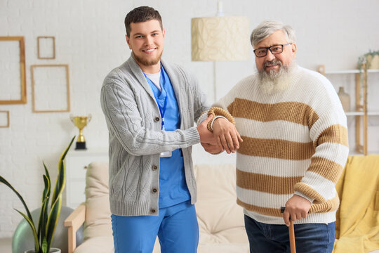 Young caregiver taking care of senior man with stick at home