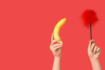 Female hands with feather stick from sex shop and banana on red background, closeup
