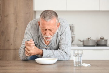 Senior man suffering from Parkinson syndrome eating soup at table in kitchen