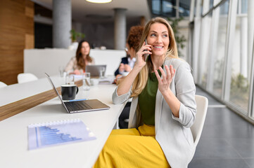 Happy businesswoman looking away and talking over mobile phone while working in meeting room with...