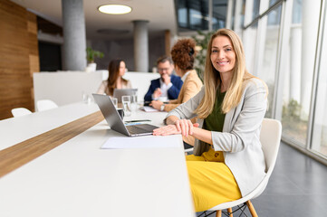 Portrait of smiling blond businesswoman working over laptop in board room while colleagues discussing in the background