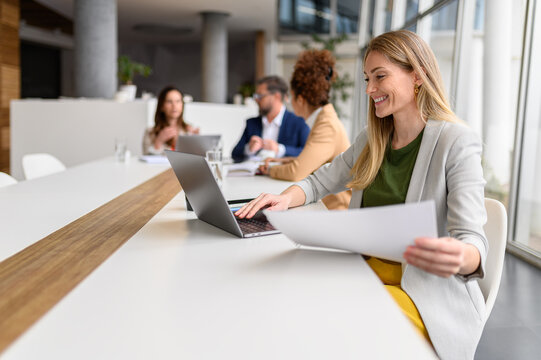 Confident businesswoman smiling and examining financial report over laptop while working at conference table