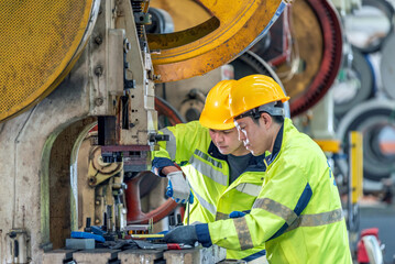 Two industrial workers wea yellow safety helmets and bright yellow reflective jackets inspecting equipment in a factory or plant environment with large machinery and pipes