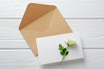 Blank invitation card, envelope and rose flower on white wooden table, flat lay