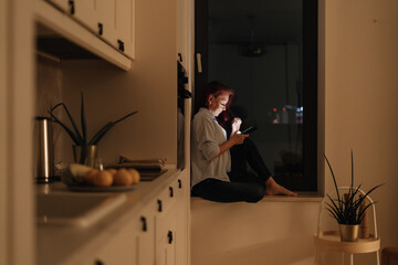 Woman sits in windowlight alone at night looking at glowing smartphone representing blue light at night digital intimacy and emotional solitude during evening scrolling routine in modern lifestyle