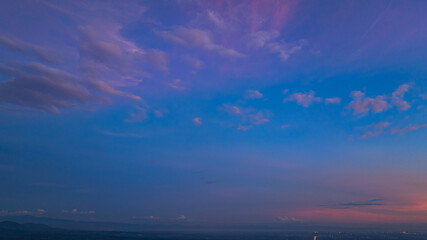 A breathtaking aerial panoramic view of a vibrant sunset illuminating the sky above a lush green hilltop with a small structure, surrounded by scattered clouds and distant landscapes.